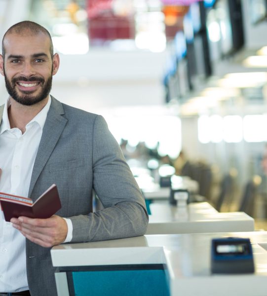 Portrait of smiling businessman standing at check-in counter with passport and boarding pass at airport terminal
